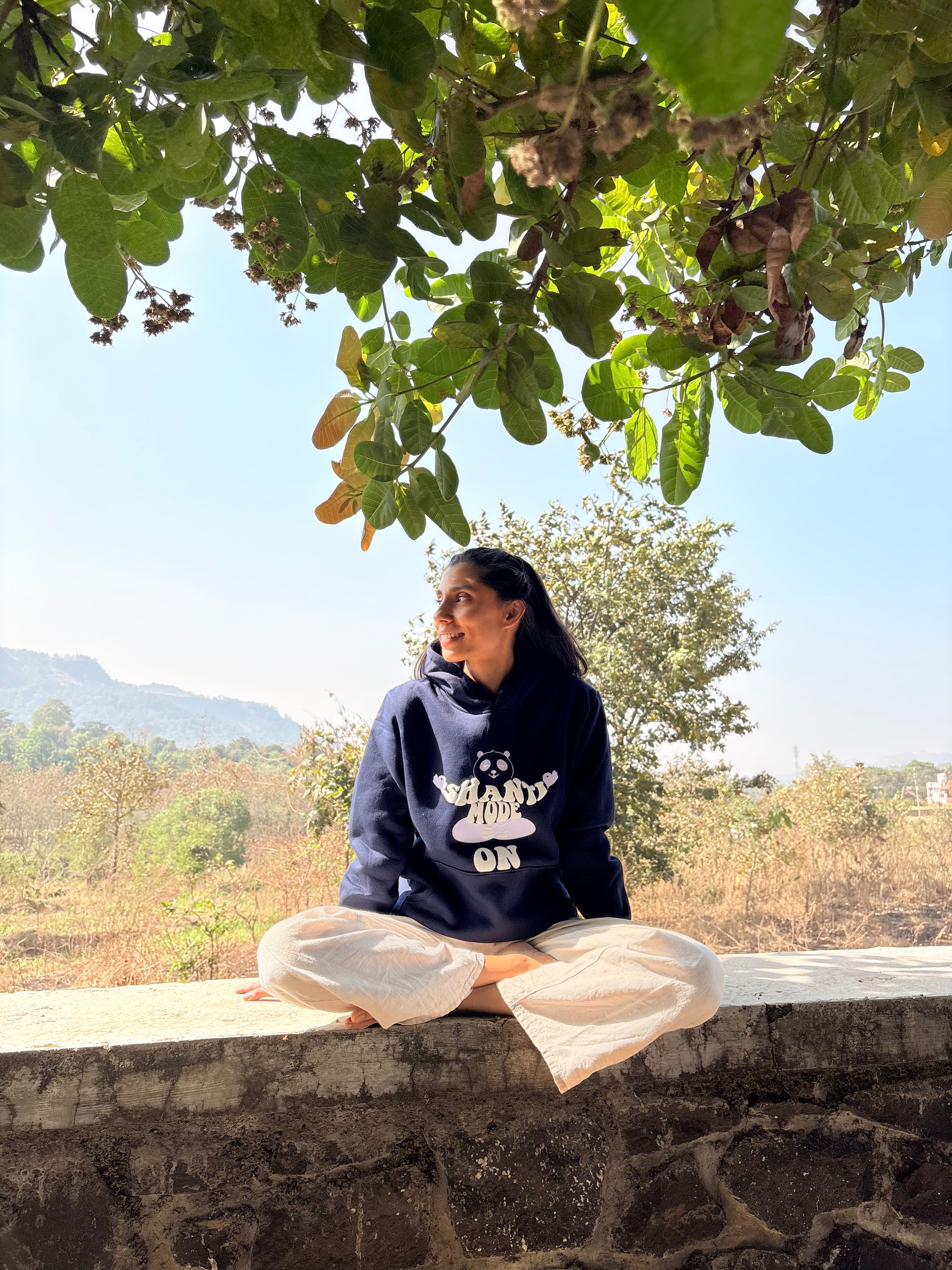 Person wearing a navy blue hoodie with text that says 'Shanti Mode On' sitting on a stone wall with greenery and mountains in the background.