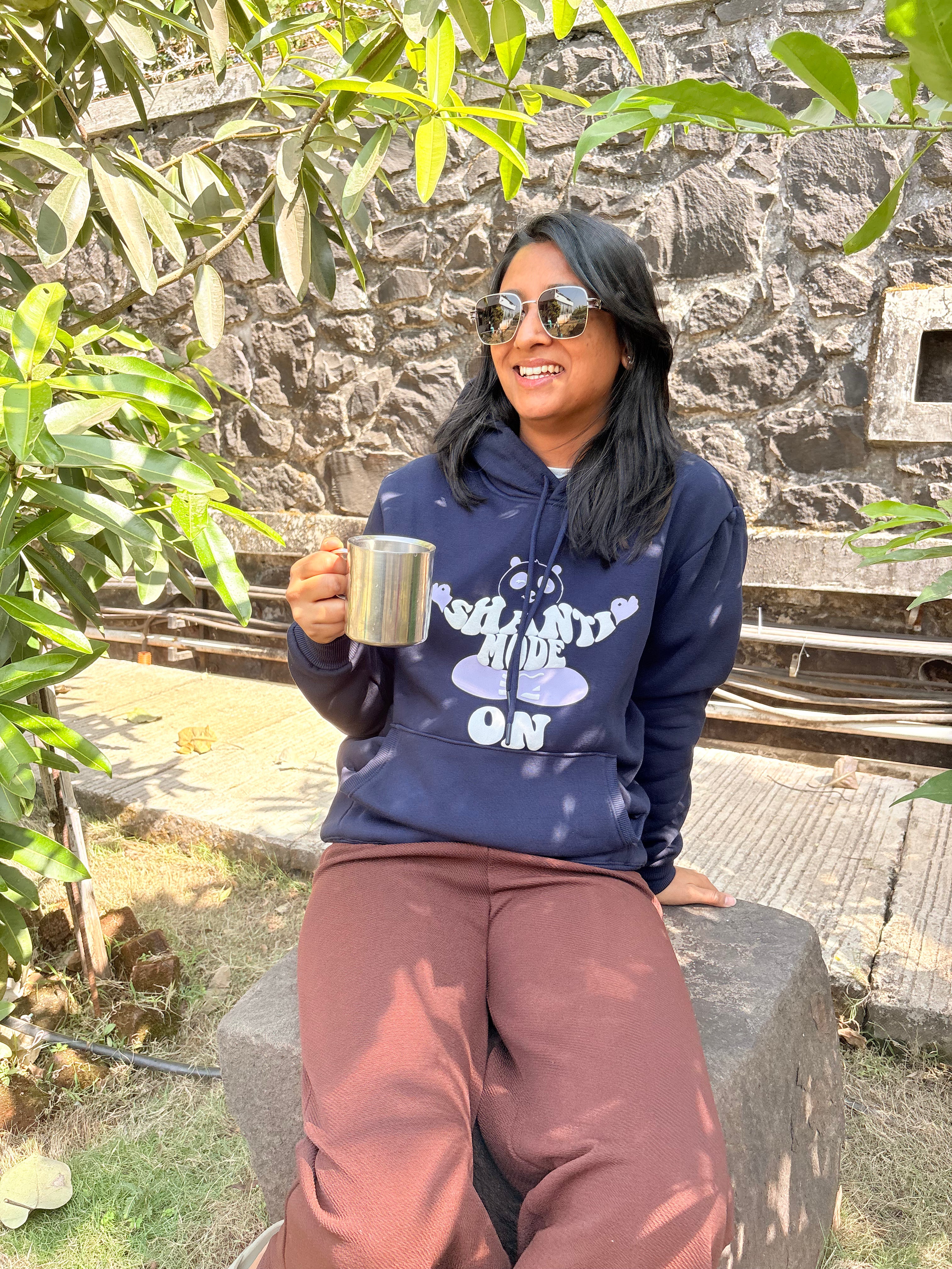 Person wearing a navy hoodie with text and graphics, sitting outdoors holding a mug.