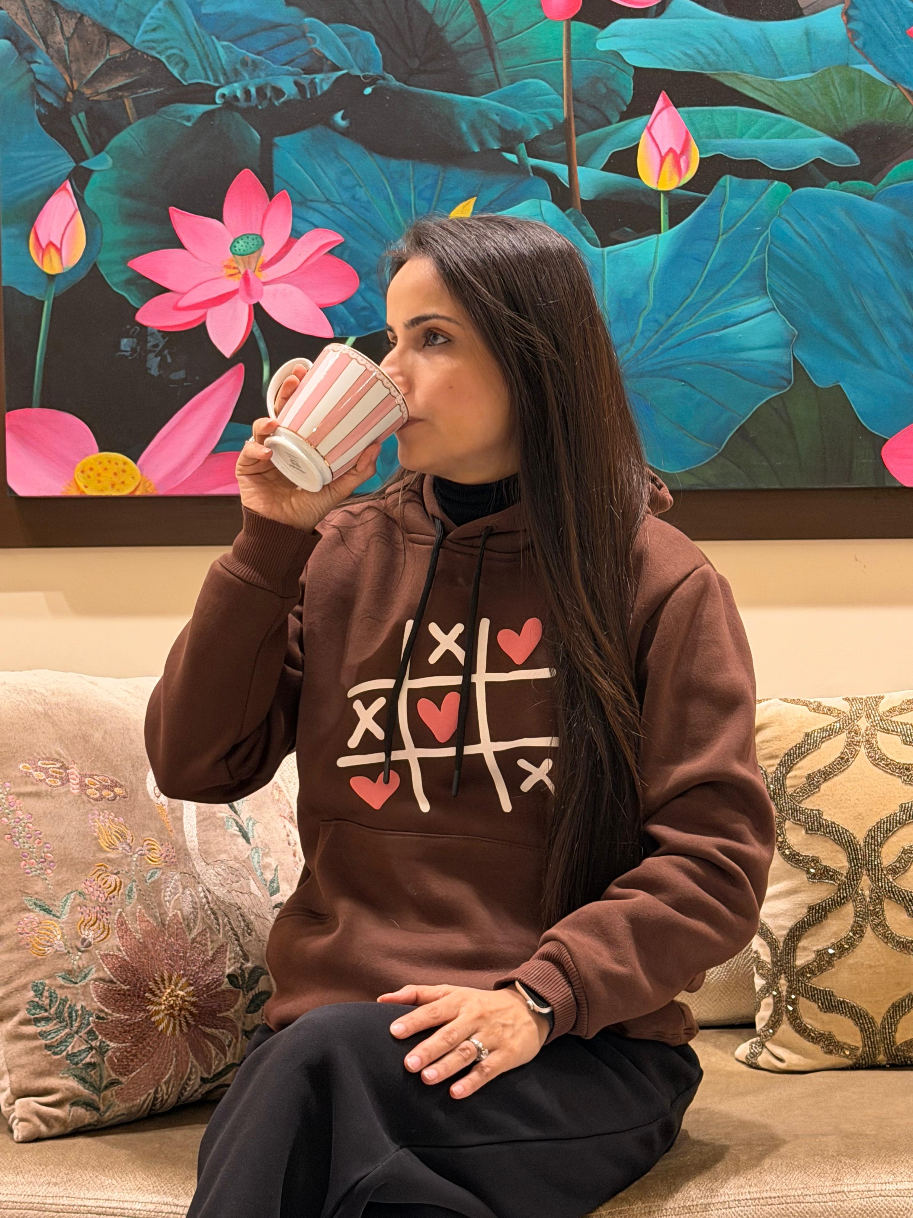 Woman wearing a brown hoodie with a tic-tac-toe design, sitting on a couch with a colorful floral painting in the background.