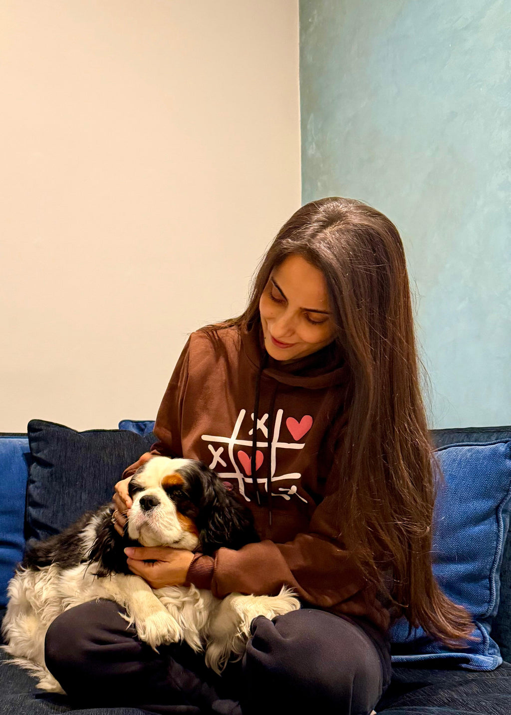 Woman sitting on a blue couch holding a dog, wearing a brown sweatshirt with heart designs.